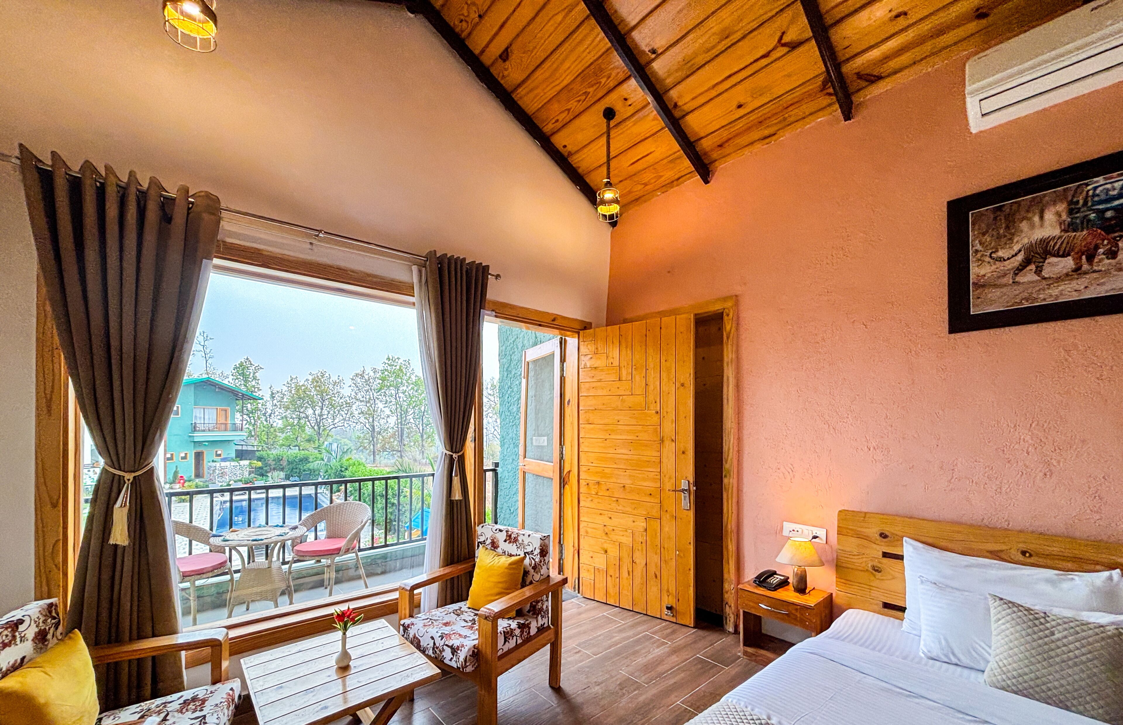 A room with a wooden ceiling, a bed with white bedding, a nightstand with a lamp, a chair with a floral cushion, and a view of a building through a window with curtains.