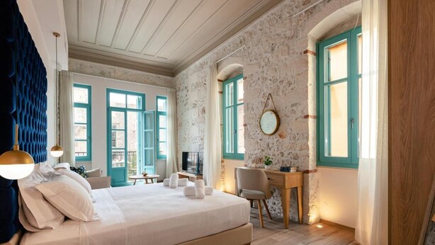 A bedroom with a large bed, a desk, and a chair. There are two windows with blue shutters and a stone wall.