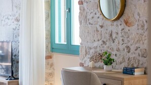 A room with a stone wall, a wooden desk, a chair, a mirror, a window with blue shutters, and a potted plant.