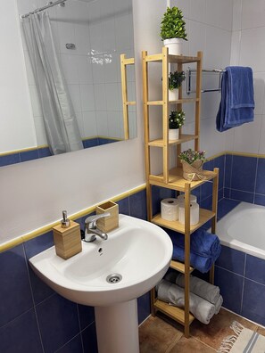 A bathroom with a white sink, a wooden shelf with plants, a blue towel, and a bathtub. - Veramarinas apartment - relax and privacy (AN)