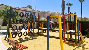 A playground with tire climbing structures and a bench, surrounded by palm trees and a building with a tiled roof. - Veramarinas apartment - relax and privacy (AN)