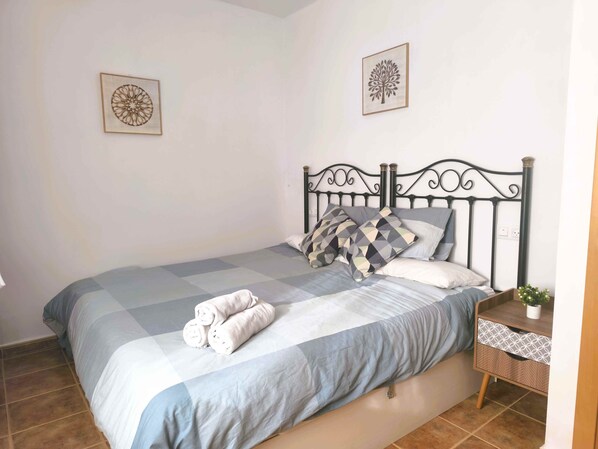 A neatly made bed with a checkered bedspread, two pillows, and rolled towels. There is a nightstand with a plant and a drawer. Two wall art pieces are visible. - Veramarinas apartment - relax and privacy (AN)