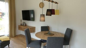Ein Essbereich mit einem runden Holztisch, sechs grauen Stühlen, einem an der Wand montierten Fernseher und einer Pendelleuchte.