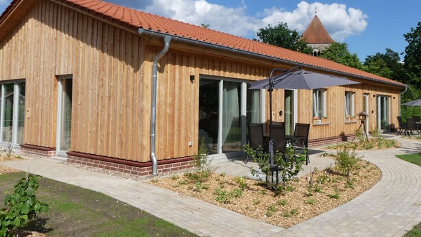 Ein Holzhaus mit rotem Ziegeldach, großen Fenstern und einer Terrasse mit Gartenmöbeln.