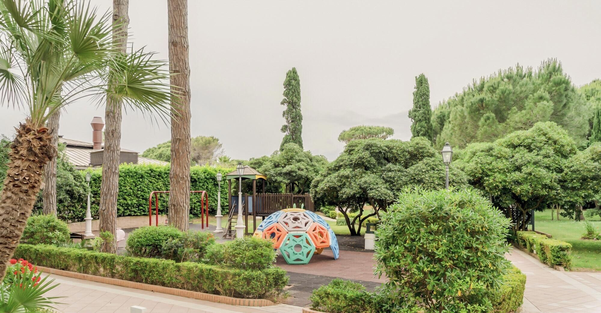 Un parque con un área de juegos, palmeras y setos bien cuidados.