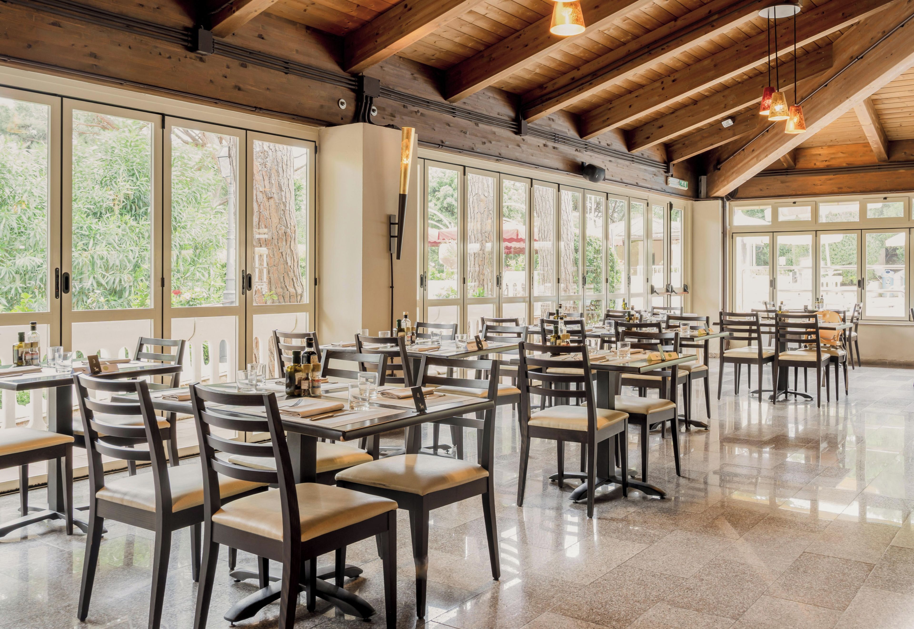 A spacious dining area with multiple tables and chairs, large windows offering views of greenery, and a high wooden ceiling with exposed beams.