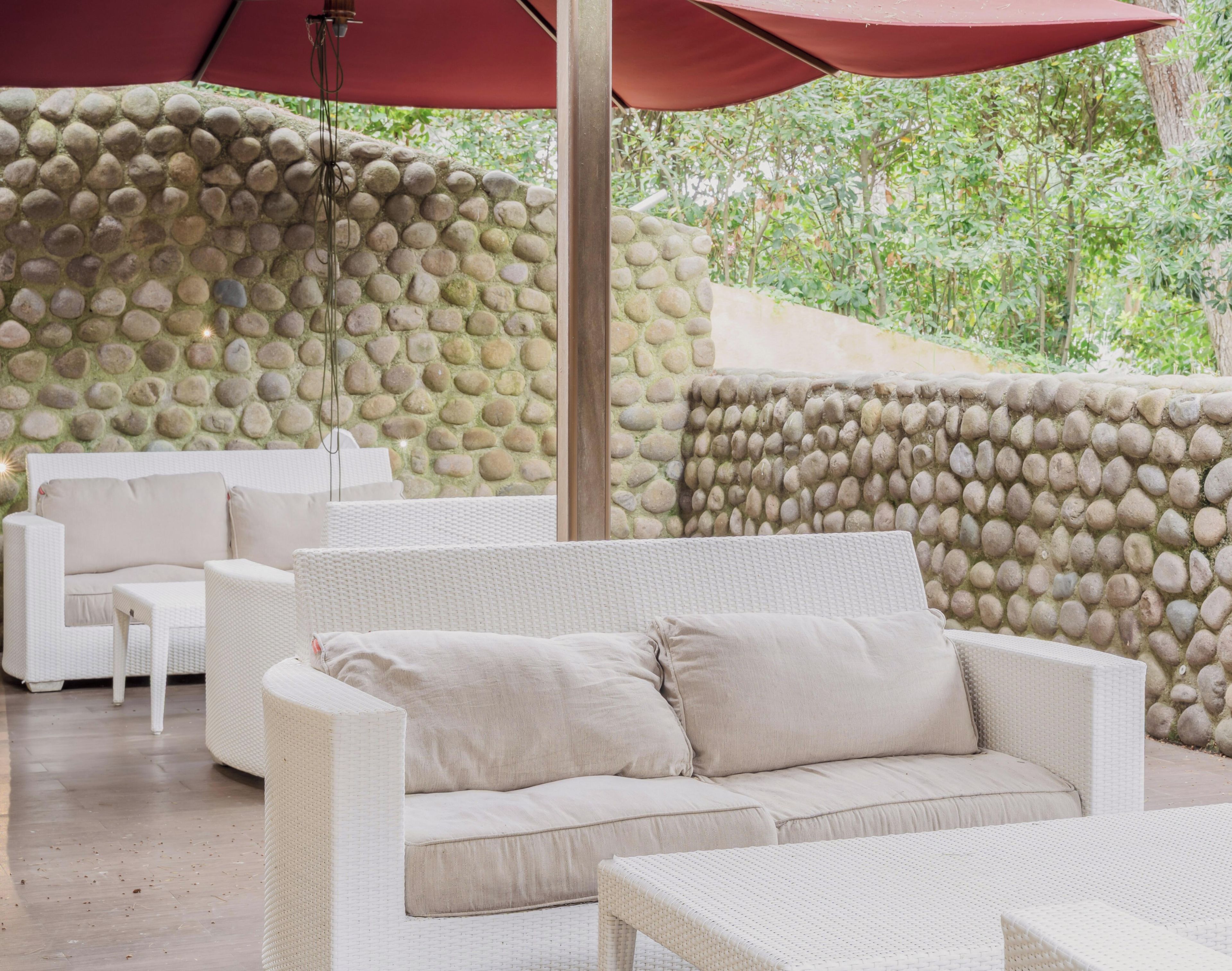A covered outdoor seating area with a stone wall, white wicker furniture, and a red umbrella.