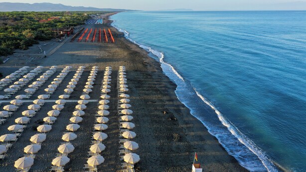A beach with rows of white umbrellas and chairs, a clear view of the ocean, and a distant shoreline with buildings. - Hotiday Marina di Bibbona (Marina di Bibbona)