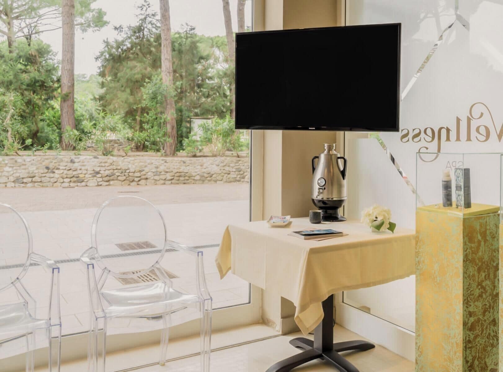 Una habitación con televisión, una mesa con cafetera, sillas transparentes y una pared de piedra vista.