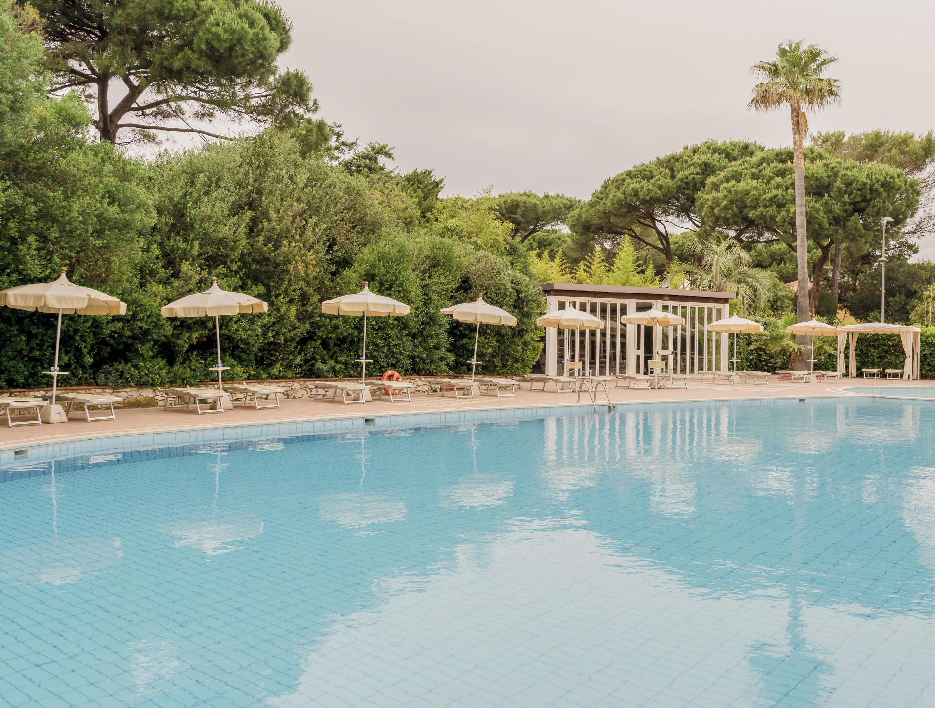 Una piscina con sillones de descanso y sombrillas, rodeada de árboles y un edificio.