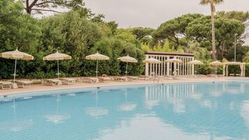 Una piscina con sillones de descanso y sombrillas, rodeada de árboles y un edificio.