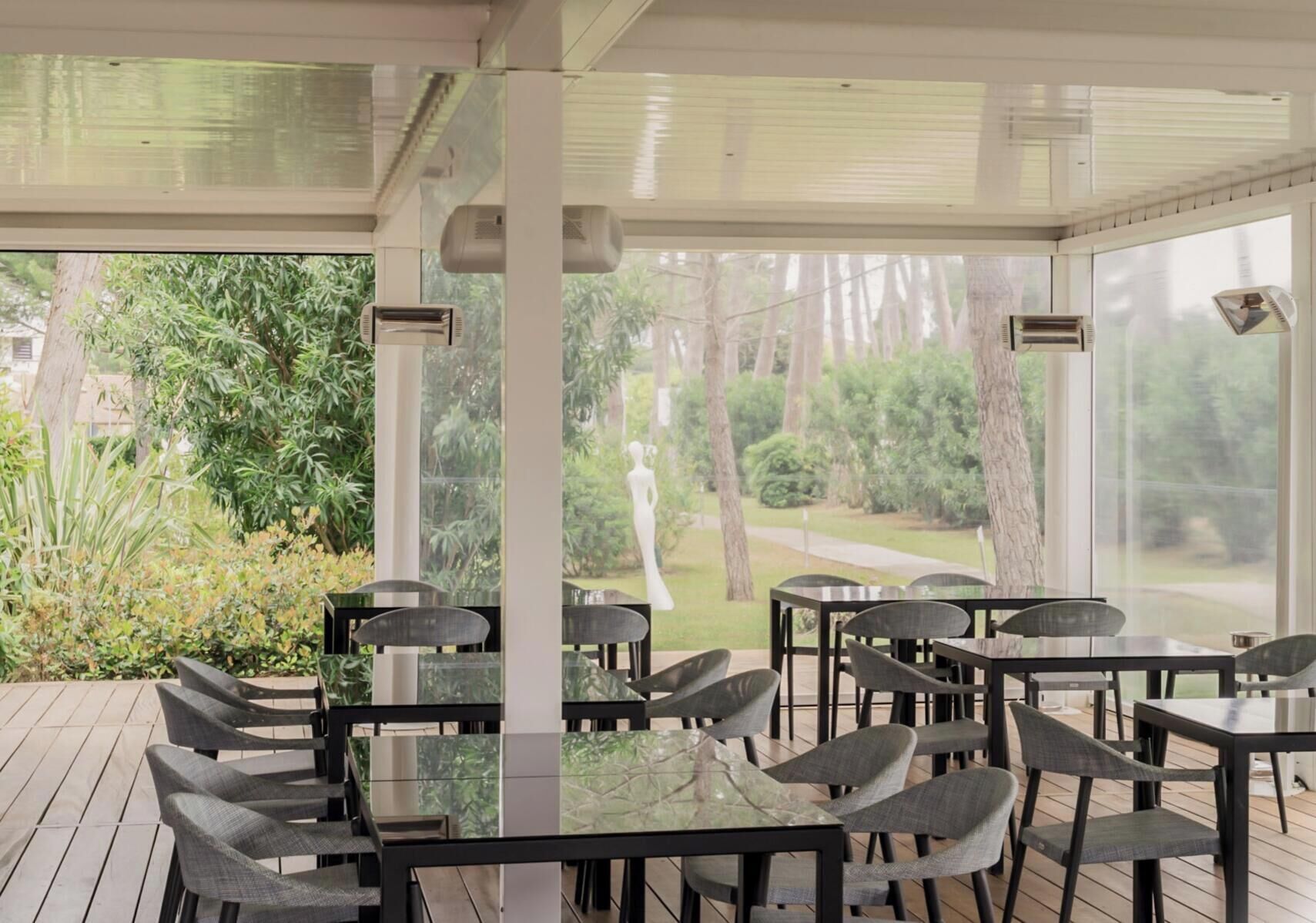 An outdoor patio with tables and chairs, surrounded by glass walls, overlooking a garden with trees and a statue.