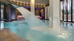 A modern indoor pool with a water slide, a lounge area with a sofa, and large windows offering a view of the outdoors. - Hotiday Marina di Bibbona (Marina di Bibbona)