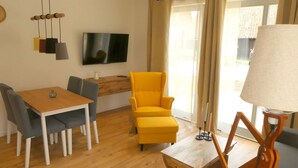 A modern living room with a wooden dining table, grey chairs, a yellow armchair, a TV mounted on the wall, and a lamp on a side table. - Alpaca Lodge Kirchwahlingen (Kirchwahlingen)