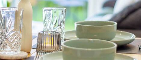 A dining table with a white vase containing a single white flower, two clear glass cups, and a set of white plates.