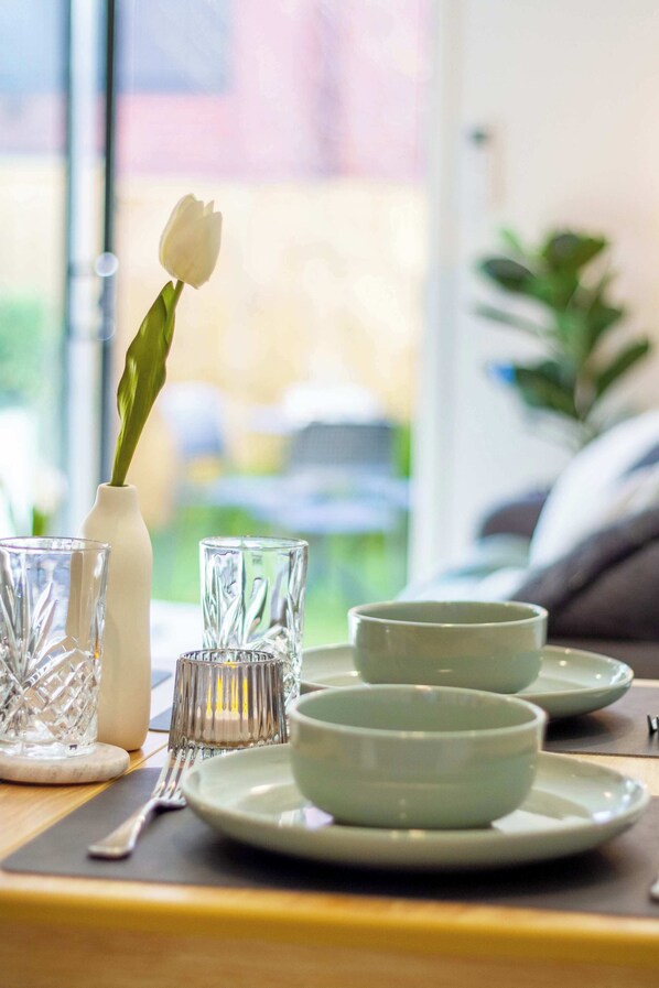 A dining table with a white vase containing a single white flower, two clear glass cups, and a set of white plates.