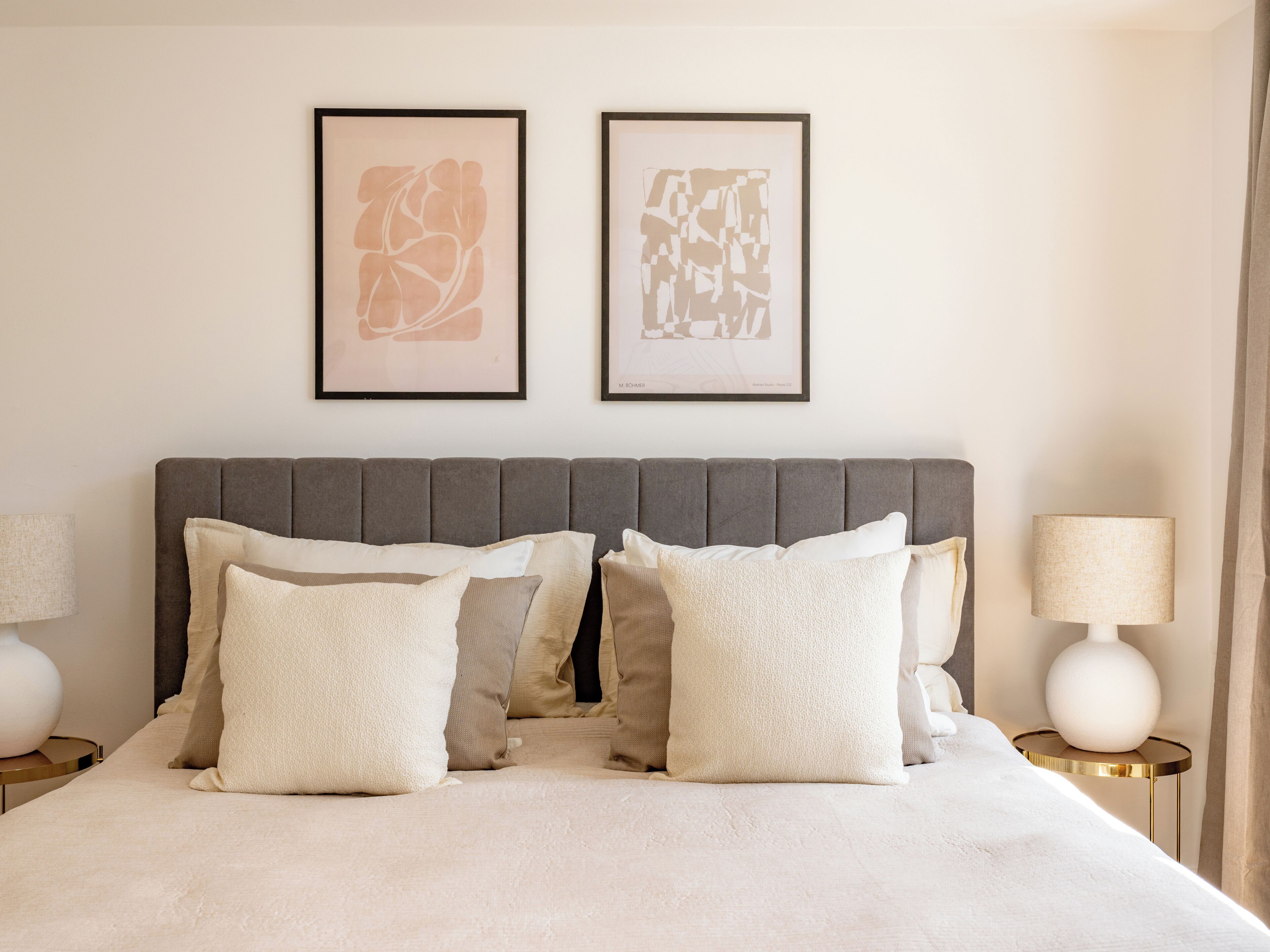 Een net opgemaakt bed met een grijs gestoffeerd hoofdbord, meerdere kussens en twee ingelijste abstracte kunstwerken aan de muur.