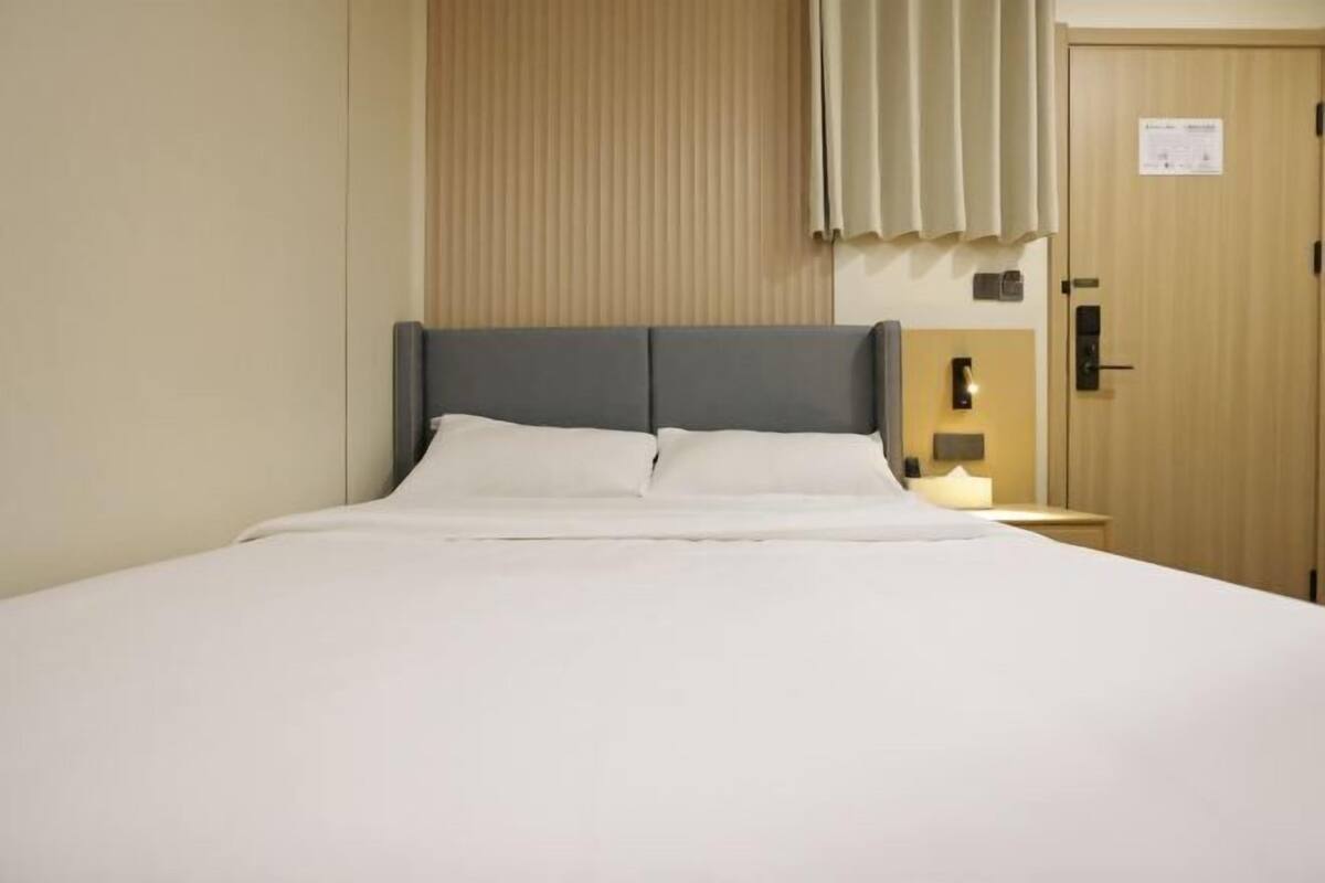 A hotel room with a bed, a nightstand, a wall-mounted lamp, and a door with a sign.