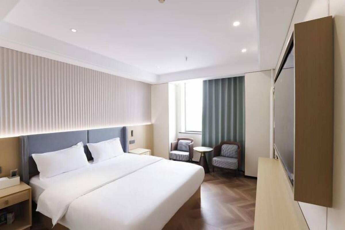 A modern hotel room with a large bed, a bedside table, a chair, a small table, and a TV mounted on the wall.