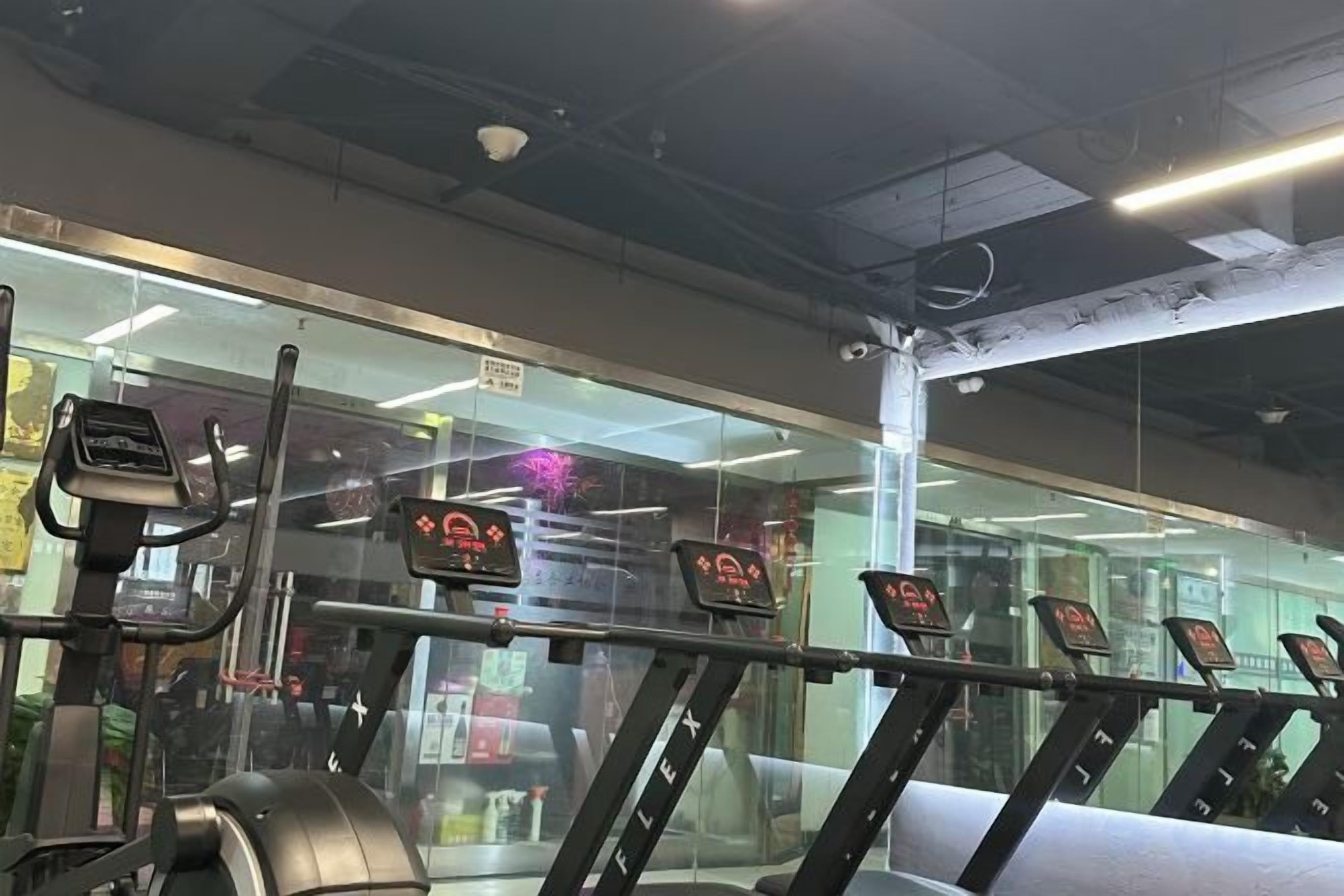 Un gimnasio con equipo de ejercicio, incluyendo cintas de correr y bicicletas estáticas, y un muro de cristal que refleja el interior.