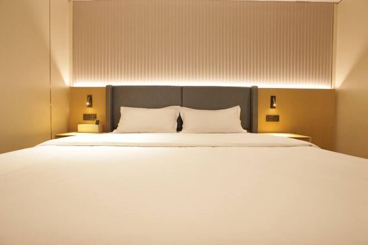 A hotel room with a large bed, a headboard with integrated lighting, bedside tables with lamps, and a window with blinds.