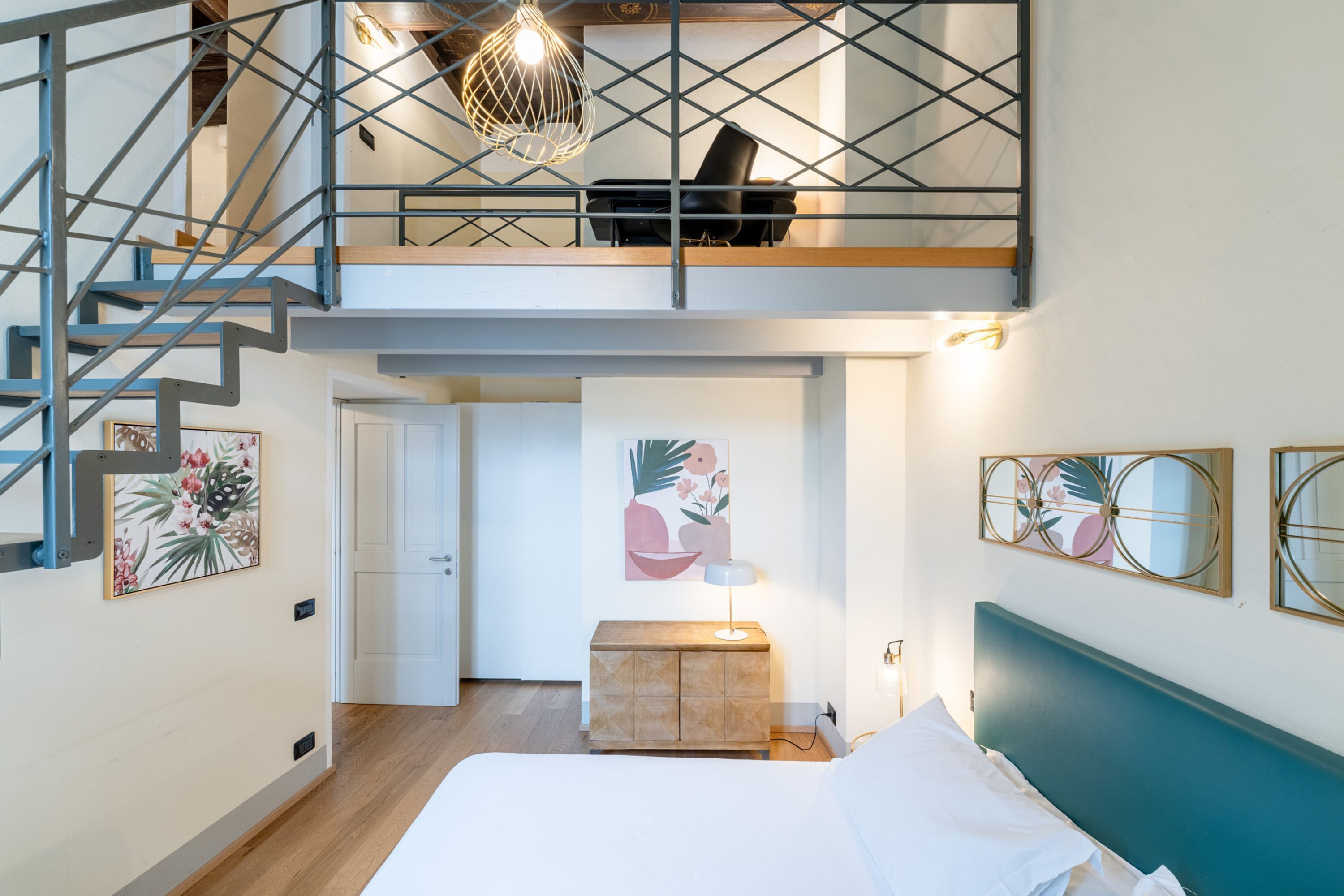 Een slaapkamer met een bed, nachtkastje, bureau, stoel, trap en kunstwerken aan de muren.