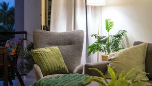 A living room with a grey armchair, a green throw pillow, a beige pillow, and a green blanket. There is a potted plant on a side table.