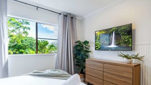A modern bedroom with a large bed, a wooden dresser, a TV mounted on the wall, and a window with a view of greenery.