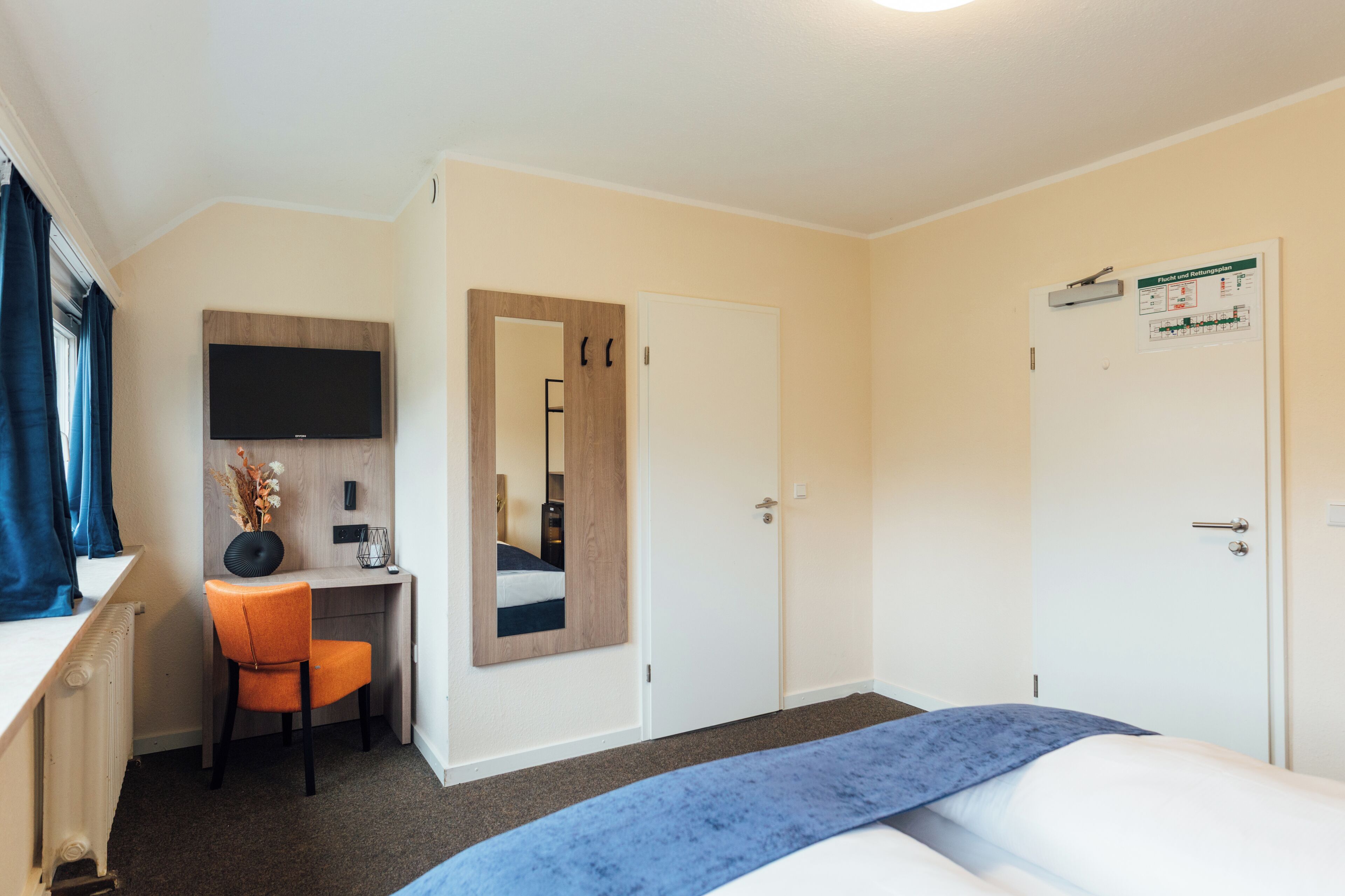 A hotel room with a bed, a desk with a chair, a television, a mirror, and a door with a bulletin board.
