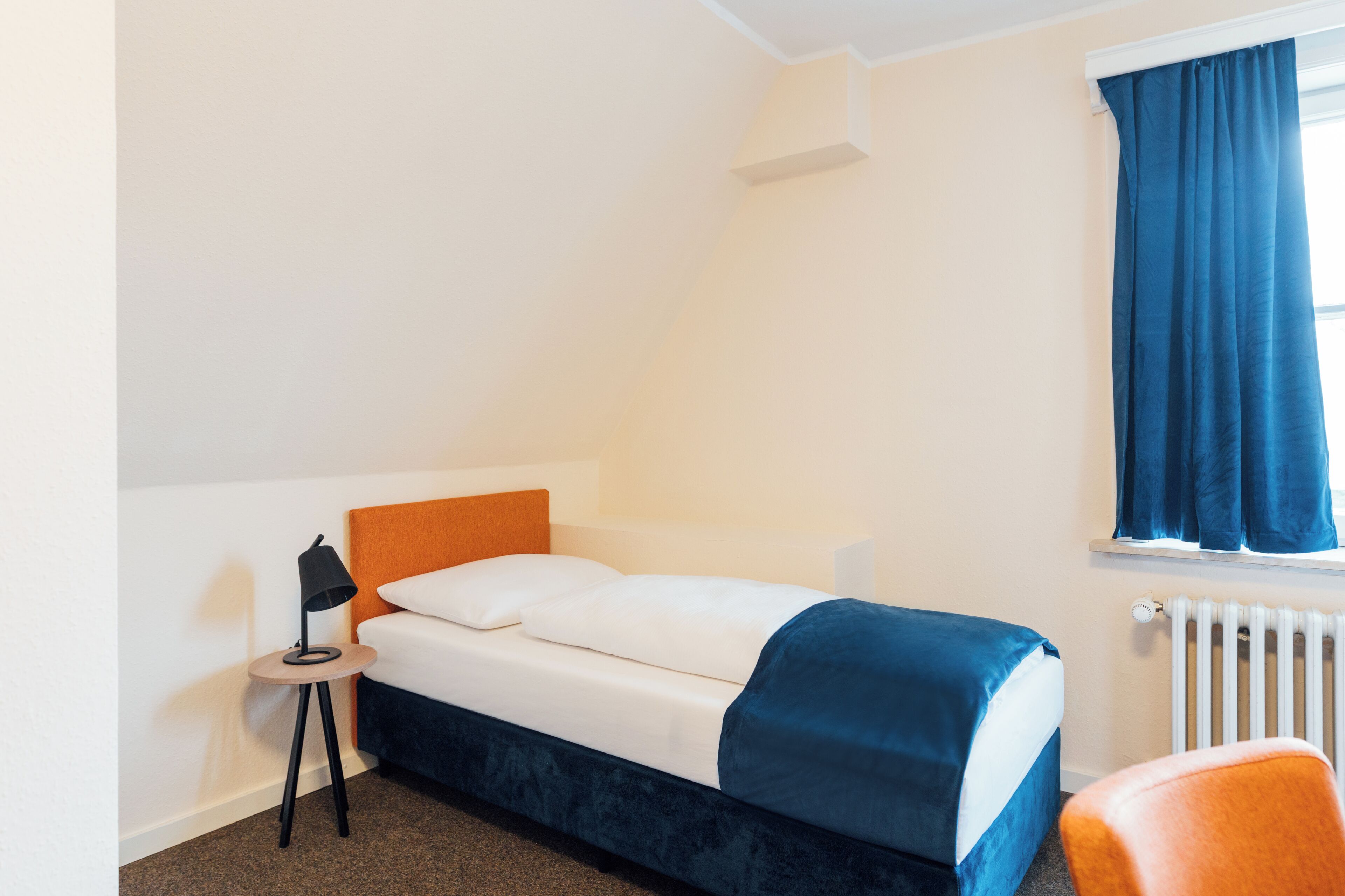 Una camera da letto con un letto, un comodino con una lampada, una finestra con tende e un termosifone.