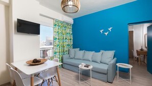 A modern living room with a blue accent wall, a grey sofa, a white dining table, and a TV mounted on the wall. - Anemi Blue apt-close to the beach (Rethymno)