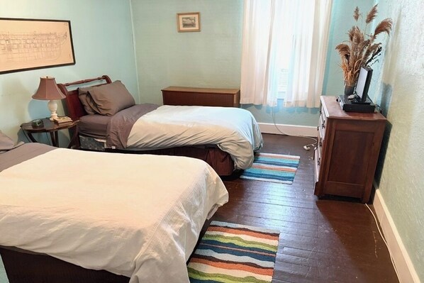 A bedroom with two beds, a nightstand, a lamp, a framed picture, a window with curtains, and a small wooden cabinet.