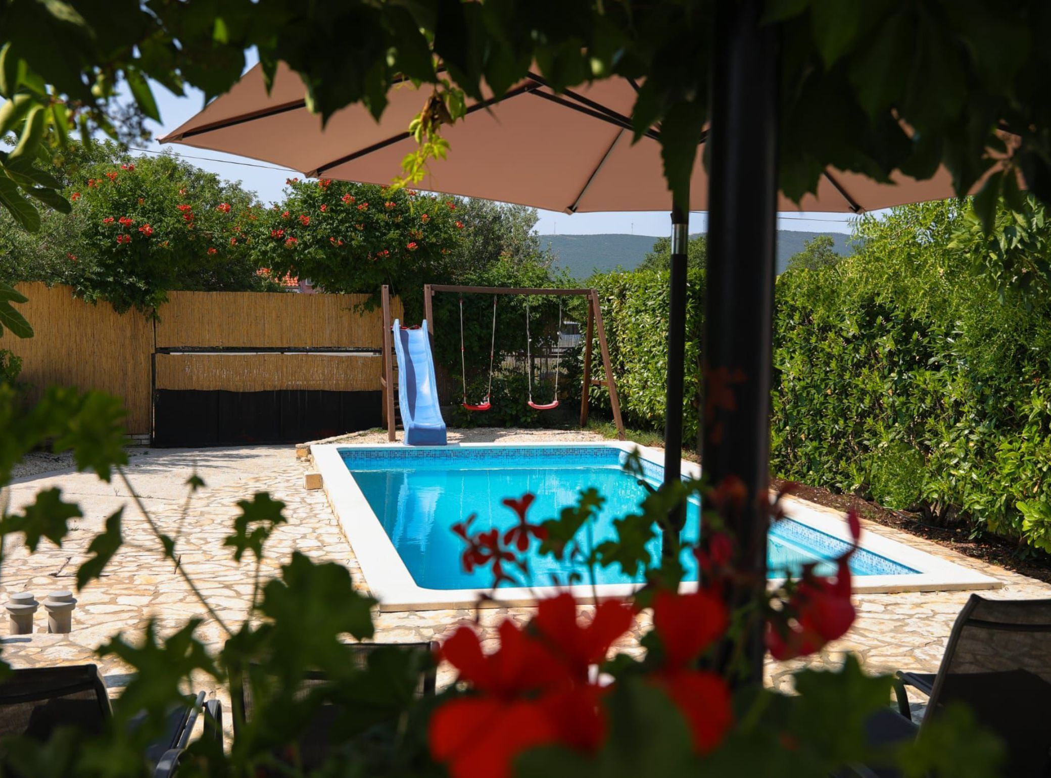 A swimming pool with a slide and swing set, surrounded by lush greenery and a wooden fence.