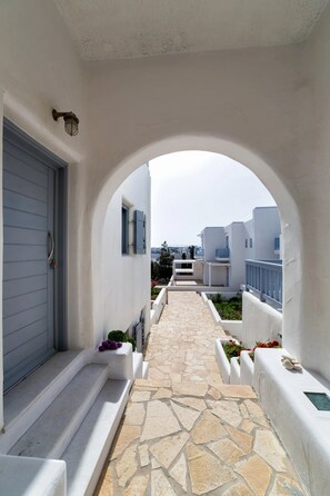 A narrow stone-paved pathway flanked by white walls and blue shutters, leading to a building with a grey door.