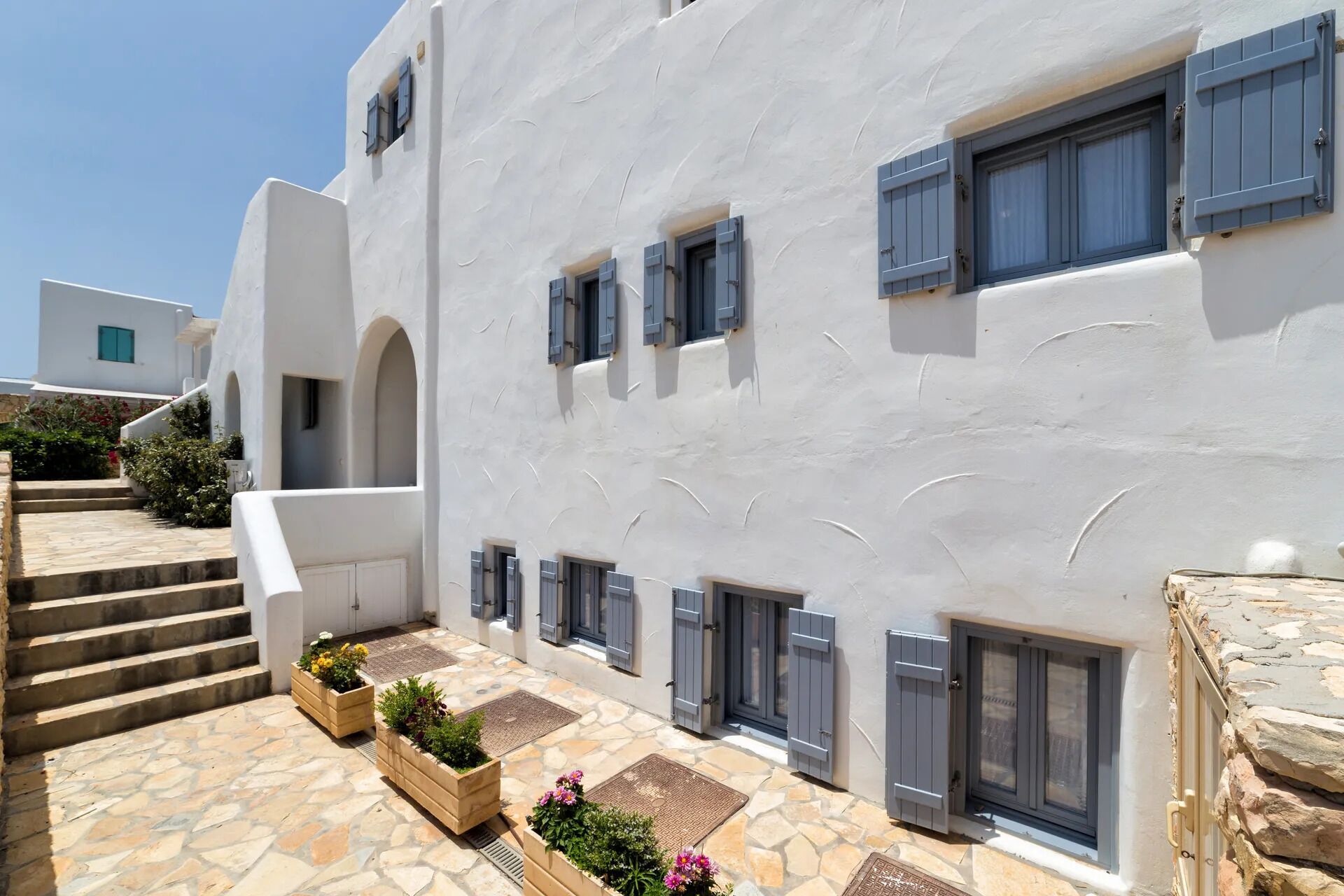 A white building with multiple windows and doors, stone steps, and wooden planters with flowers.