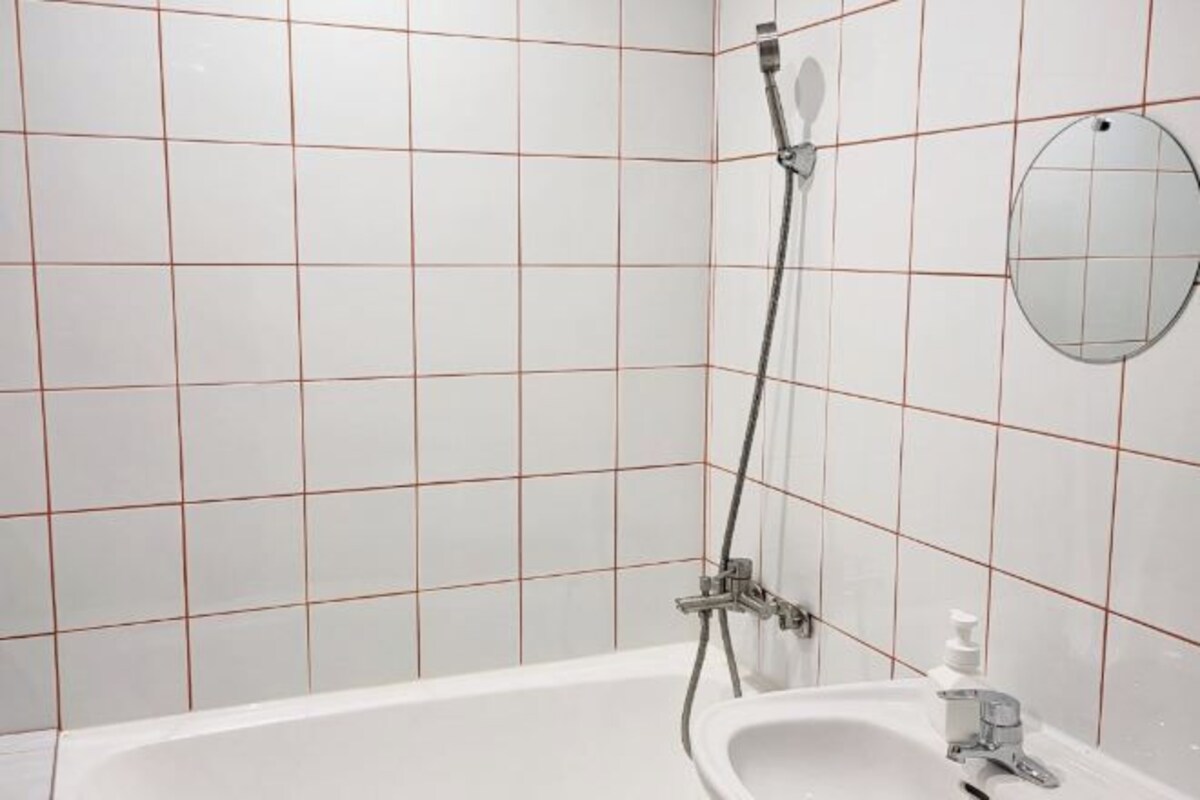 Uma casa de banho com uma banheira branca, um chuveiro manual, um espelho redondo e uma pia com dispensador de sabonete.