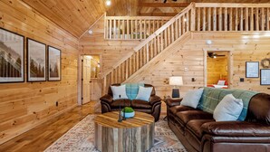 L’intérieur d’un chalet en bois est aménagé avec un escalier, des canapés en cuir, une table basse ronde et des tableaux encadrés qui ornent les murs.