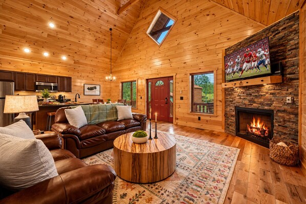 A cozy cabin interior with a fireplace, leather sofa, wooden dining table, and a kitchen area.