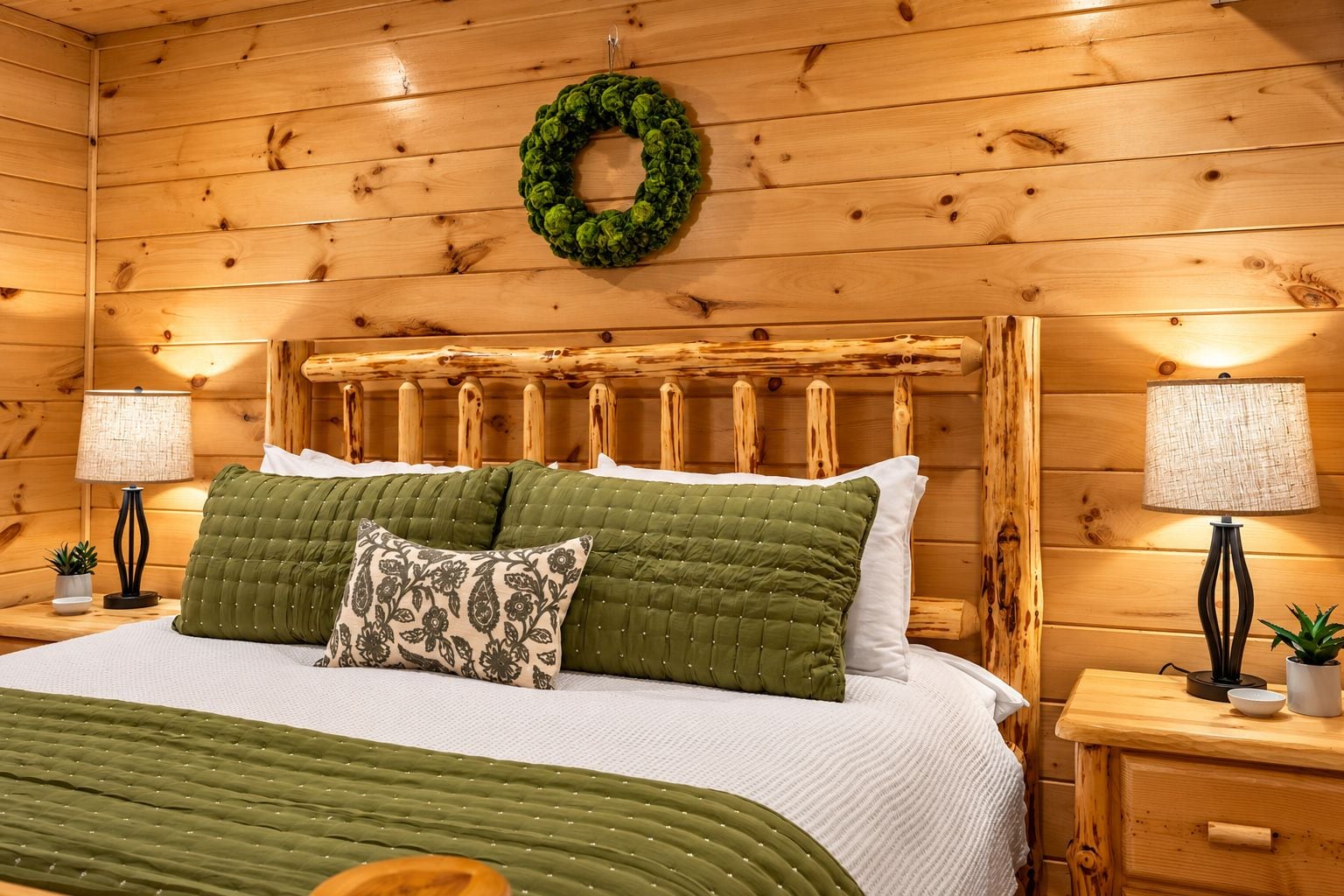 A wooden-paneled room with a bed, two bedside lamps, a green wreath on the wall, and a small plant on the nightstand.