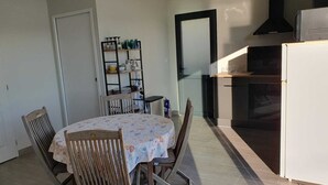 Zona pranzo con un tavolo rotondo e quattro sedie di legno, un piccolo ripiano con oggetti da cucina e un angolo cottura con piano cottura e forno.