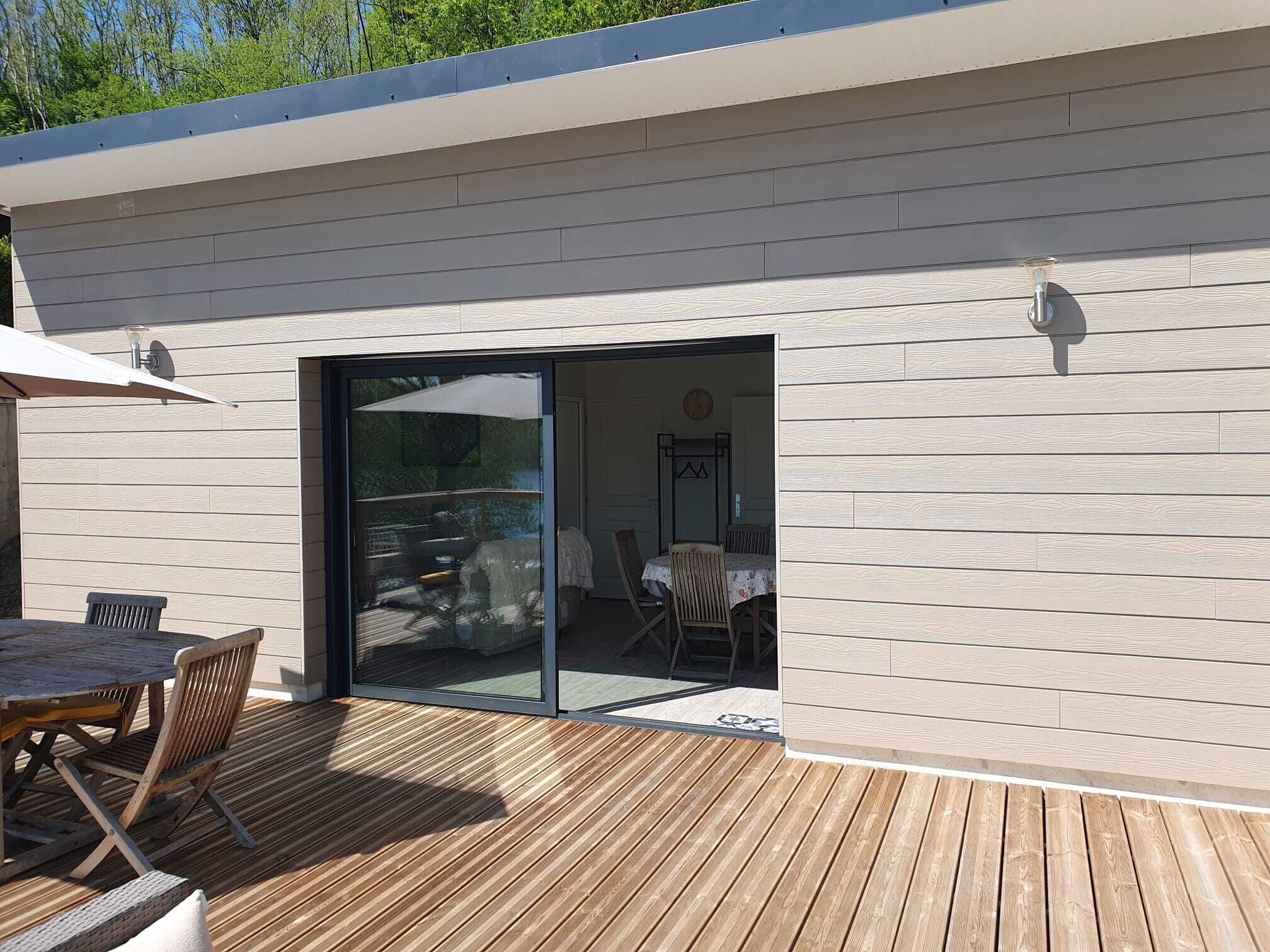 A modern outdoor patio with a wooden deck, a table with chairs, and a glass door leading to an interior space with a dining table and chairs.