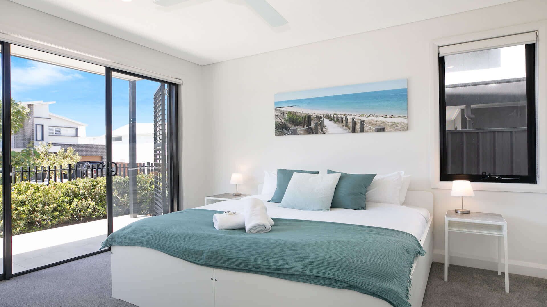 Une chambre moderne avec un grand lit, deux tables de chevet, un tableau représentant une plage et une vue sur l’extérieur grâce à une porte coulissante.