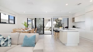 A modern kitchen with a central island, a dining area with a table and chairs, and a living room with a sofa and cushions.