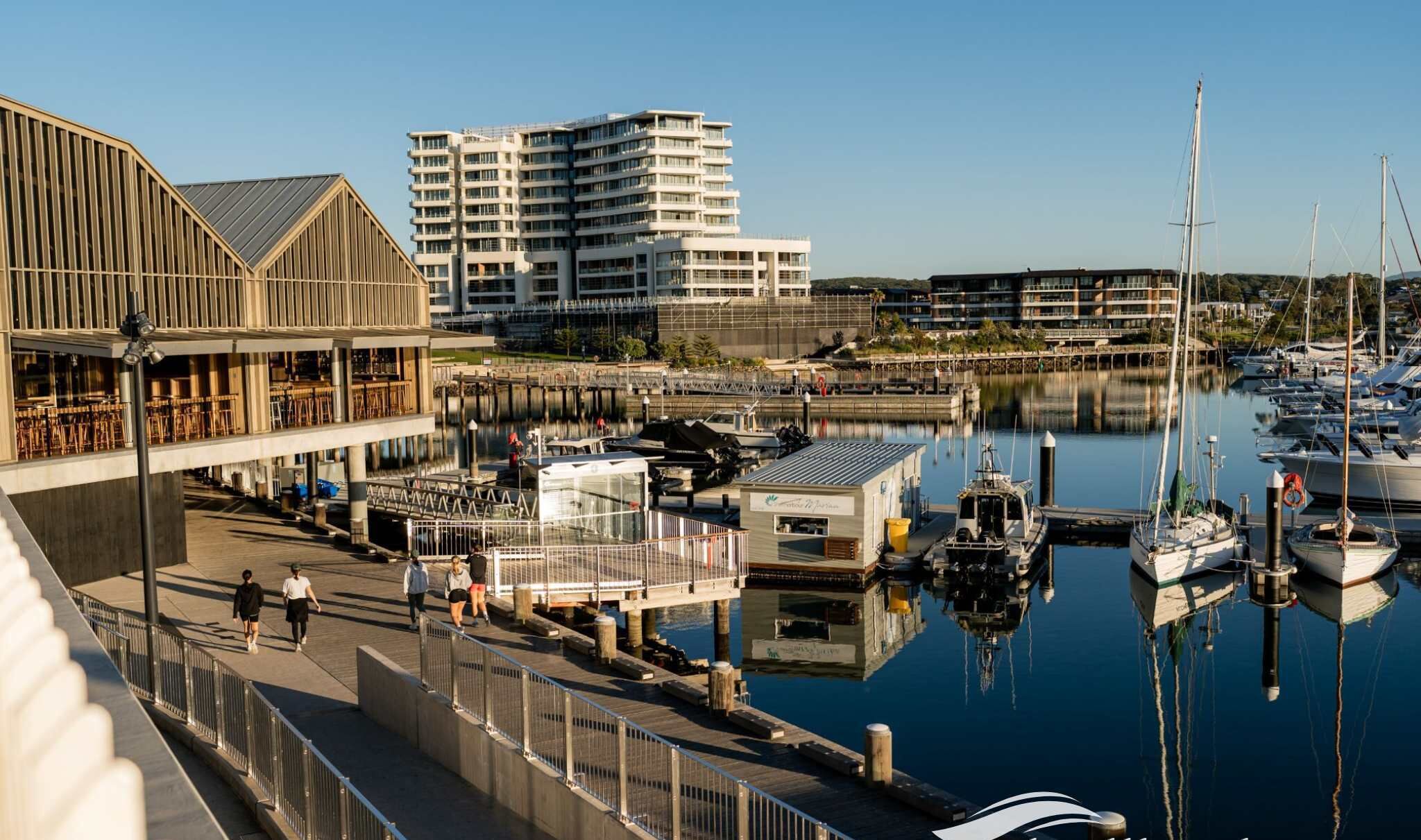 A marina with boats docked, a waterfront restaurant, and modern buildings in the background.