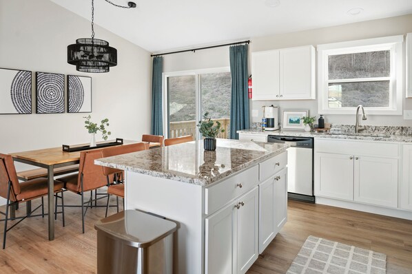 A modern kitchen with white cabinets, a granite countertop, a dining area with a wooden table and chairs, and a large window with curtains. - NEW! Charming 3-bedroom home with WiFi, near Ark Encounter, King Bed, sleeps 7. (Dry Ridge)