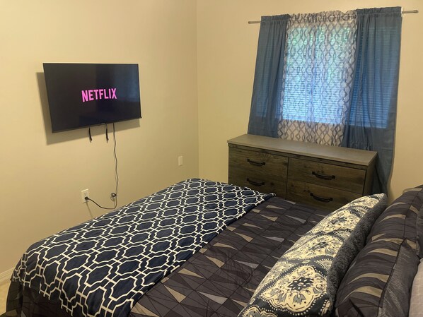 Une chambre à coucher comprenant un lit, une commode, un téléviseur fixé au mur et une fenêtre agrémentée de rideaux à motifs.