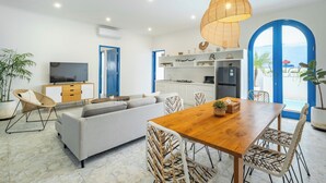 A modern living room with a sofa, dining table, and chairs, a kitchen area with white cabinets, and a large TV. - Casa Posa Seminyak (Seminyak)