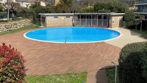A swimming pool with a wooden deck, surrounded by greenery and a stone wall.