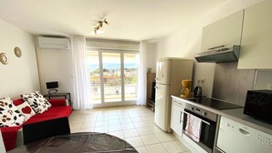 A compact living space with a kitchenette, a sofa with patterned cushions, a dining table, and a view through a sliding door to an outdoor area.