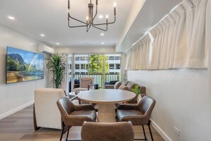 A modern dining area with a round table, six chairs, a chandelier, and a large window with curtains.